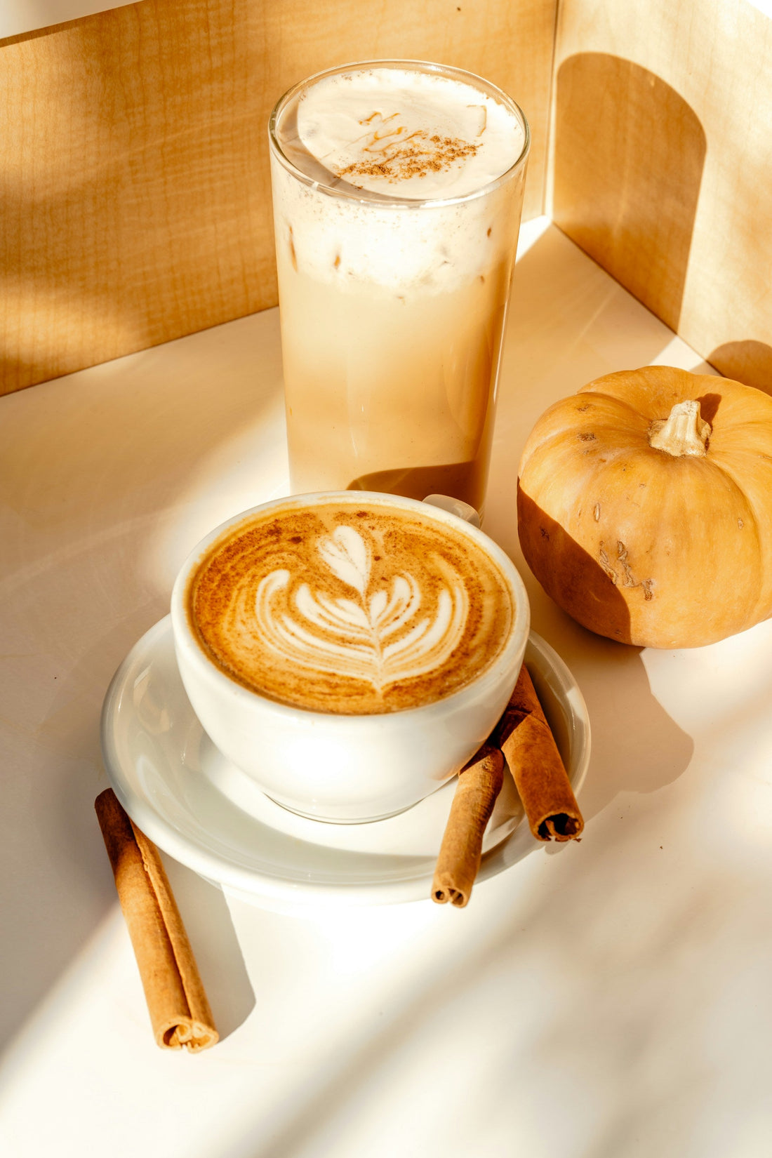 Hot latte with leaf-shaped foam art and an iced latte beside a small pumpkin and cinnamon sticks in warm sunlight.
