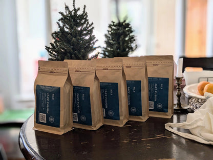 Five coffee bags from Octave Coffee Co. labeled "No.9 Jazz Fusion" are neatly lined up on a wooden table. The bags are standing upright with their blue labels facing forward, displaying key details like the roast name, logo, and a QR code. In the background, there are two small Christmas trees, a candleholder, and a basket of oranges on the table, creating a cozy, holiday-themed setting.