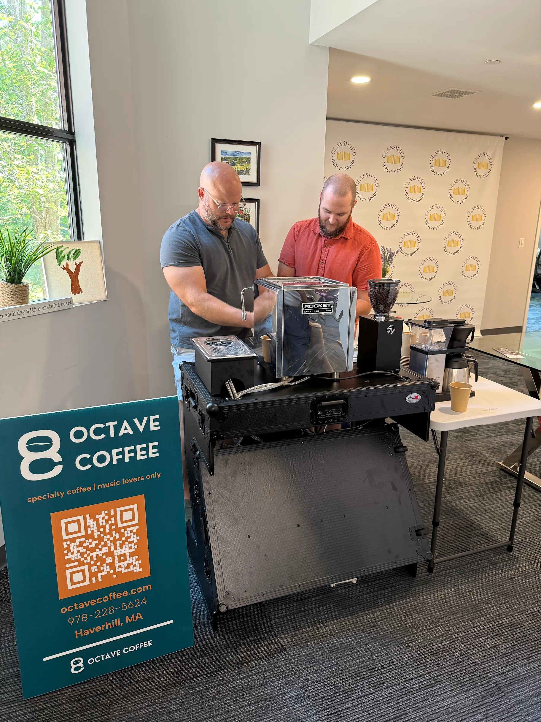 Two men prepare espresso drinks at a mobile coffee cart setup indoors. One man in a gray shirt operates a Rocket espresso machine while the other man in a red shirt stands beside him. A teal sign with the Octave Coffee logo, QR code, contact information, and location is displayed to the left of the cart. The setup includes a grinder and accessories on a small folding table, with a backdrop of a bright window and a branded wall in the background.